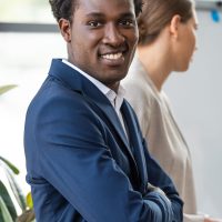 smiling african american businessman standing with crossed arms near colleague and looking at camera